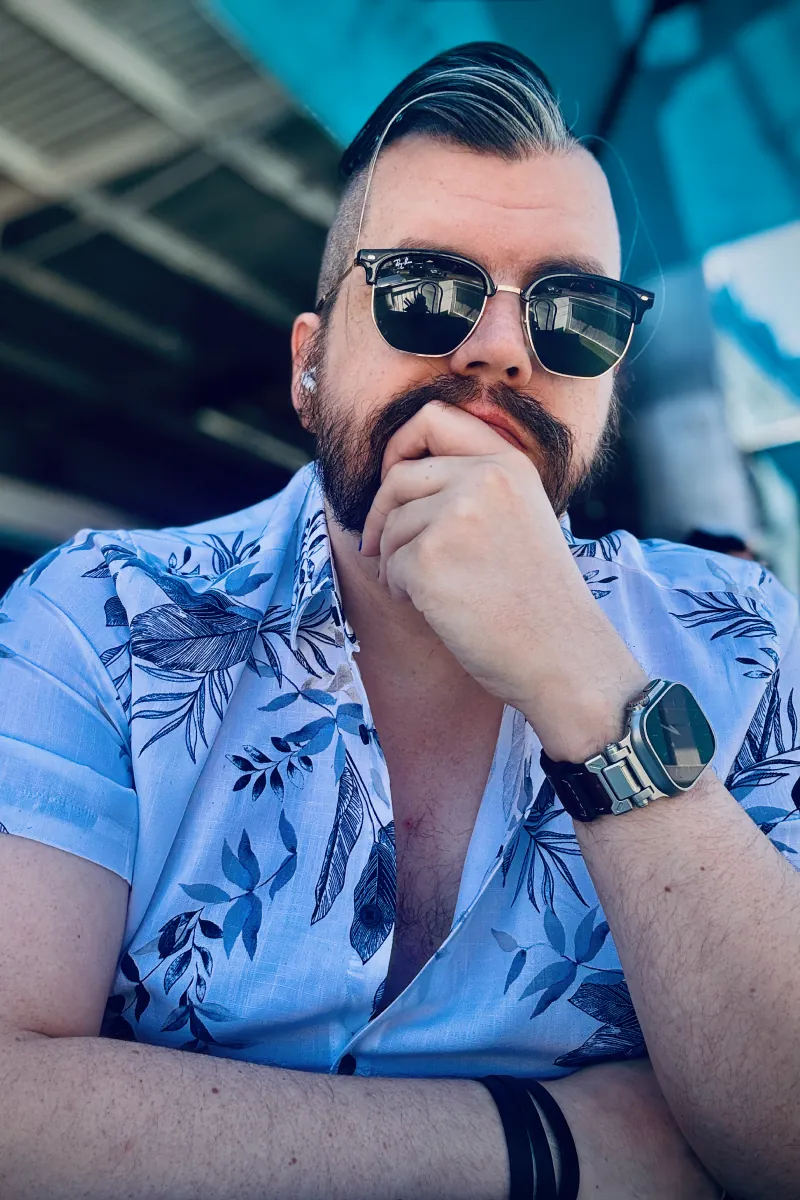 Photo of me, Jono. I'm sitting at a table in a white shirt with a floral pattern, resting my chin on my left hand. I've got a beard and slicked-back brown hair with a grey streak on the front-left. I'm wearing Ray-Ban clubmaster style sunglasses, an Apple Watch Ultra with a metal and leather band on one wrist, and a thin leather strap wrapped around the other wrist.